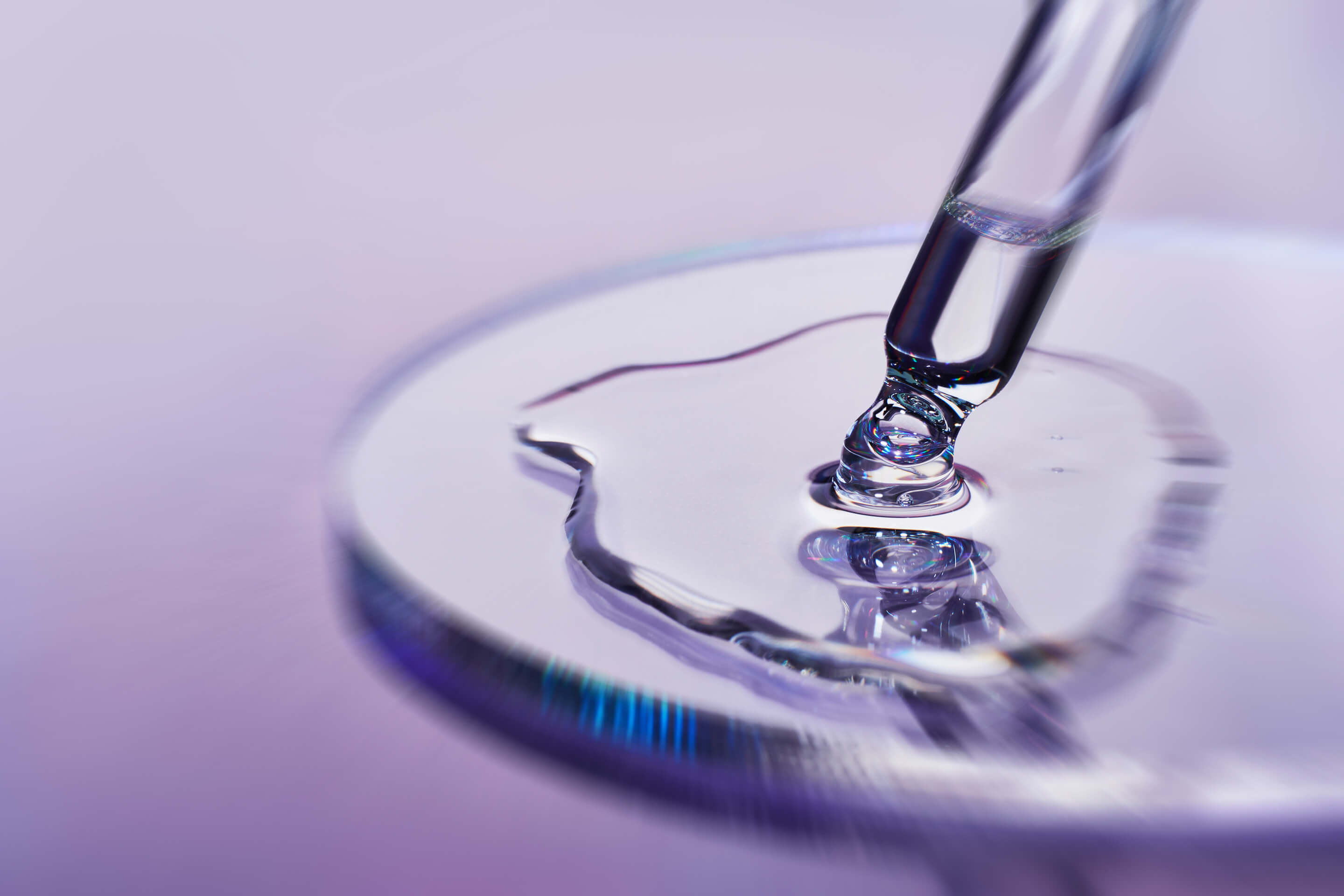 Close-up of a dropper adding liquid to a round, purple surface with a blurred purple background.